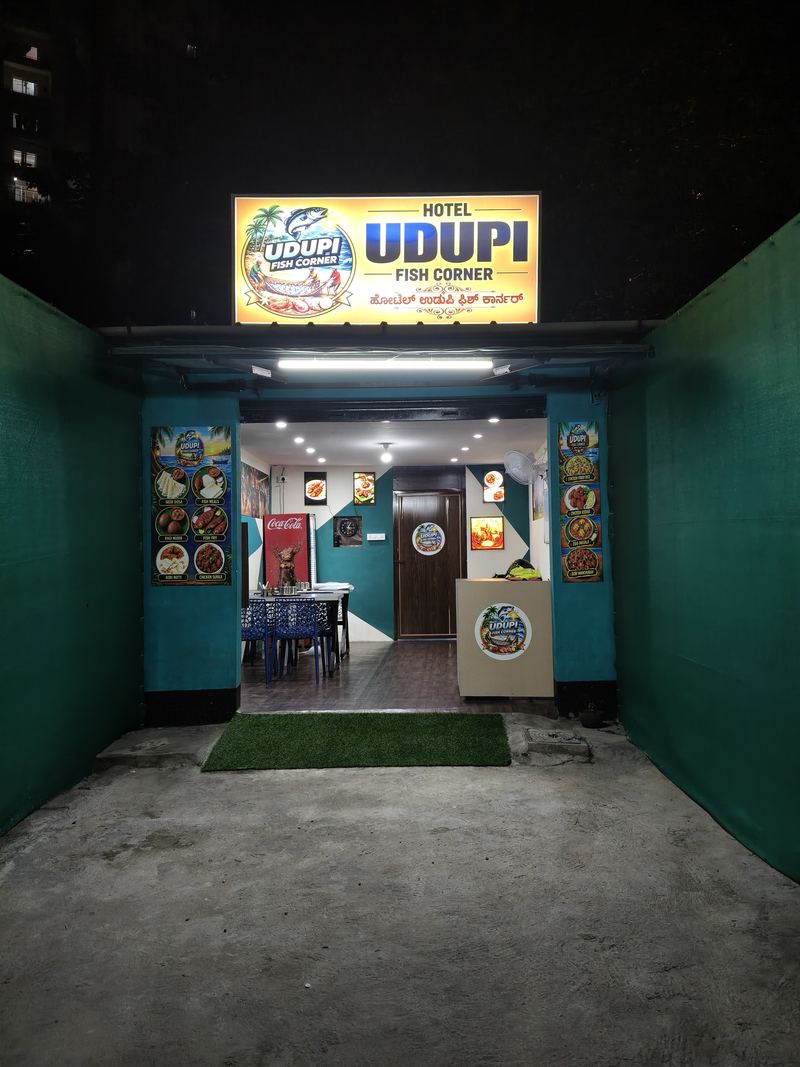 Front of Hotel Udupi Fish Corner at night: the lit sign reads Hotel Udupi Fish Corner in English and Kannada above the entrance